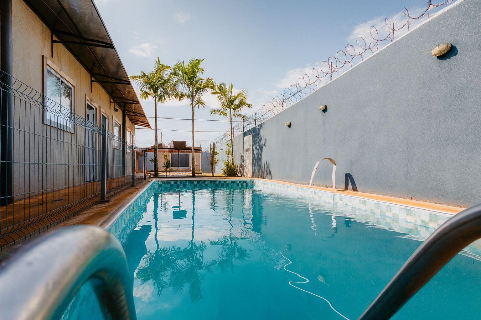A swimming pool with a metal railing, surrounded by a fence and palm trees.