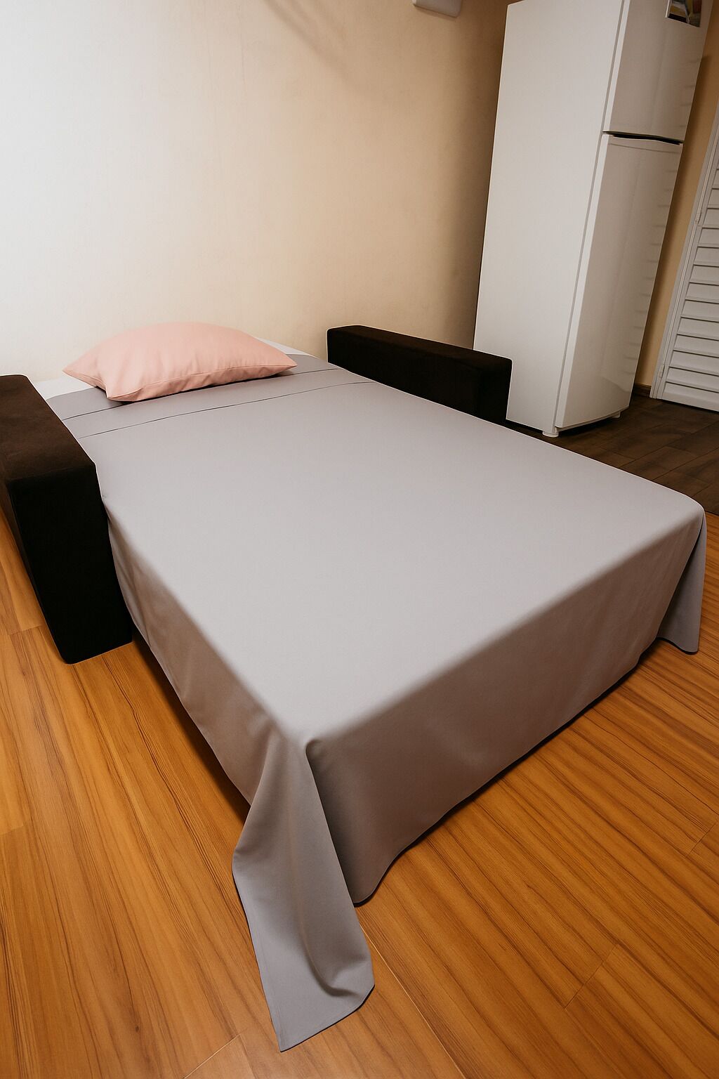 A single bed with a grey sheet, a black bench, and a white wardrobe in a room with wooden flooring.