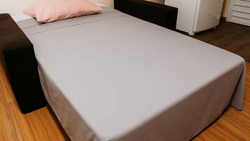 A single bed with a grey sheet, a black bench, and a white wardrobe in a room with wooden flooring.