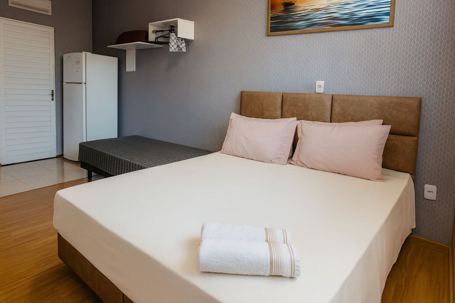 A neatly made bed with a white comforter and two pillows, a small bedside table with a lamp, and a wall-mounted shelf above the bed.