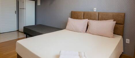 A neatly made bed with a white comforter and two pillows, a small bedside table with a lamp, and a wall-mounted shelf above the bed.