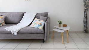 Ein graues Sofa mit weißer Decke und blumigen Kissen, ein kleiner weißer Nachttisch mit Pflanze und ein runder Spiegel an der Wand.