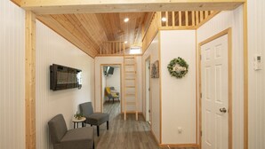 A hallway with a wooden ceiling, a TV mounted on the wall, a door with a wreath, and a chair with a small table. - Modern Cottage Cabin, private area with pond, fishing nearby, swimming and more  (McGregor)