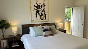Ein ordentlich eingerichtetes Schlafzimmer mit einem großen Bett, einem Wandbild, Nachttischlampen, einem Blumentopf und einer Tür zu einem Außenbereich.