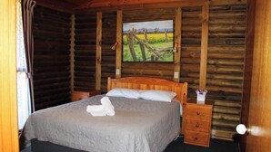 A log cabin-style bedroom with a wooden bed, a nightstand, a dresser, and a painting on the wall. - Cosy cabin in the Grampians with mountain views (Wartook)