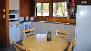A compact kitchen with wooden cabinets, a microwave, a stove, and a sink. There's a wooden table with chairs and a bottle on it. - Cosy cabin in the Grampians with mountain views (Wartook)