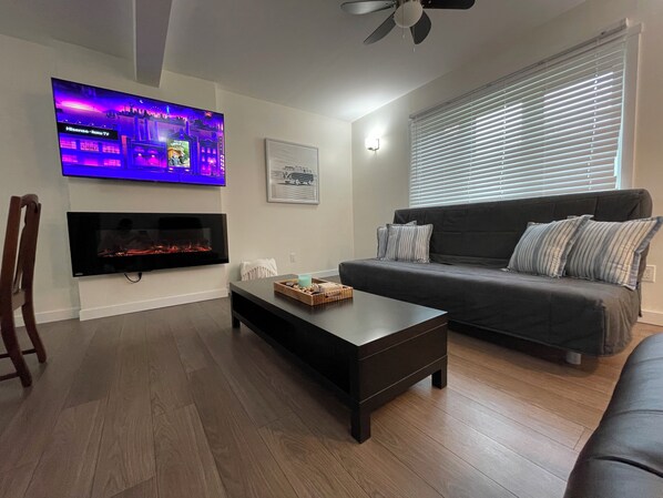 A living room with a flat-screen TV mounted on the wall, a fireplace below it, a dark-colored sofa with striped pillows, a wooden coffee table, and a ceiling fan. - Cabin in South Beach - Steps from the beach (Gimli)