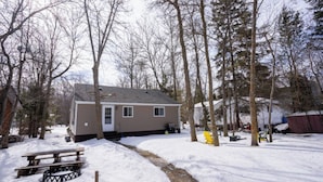 A single-story house with a front porch, surrounded by snow-covered ground and trees. - Cabin in South Beach - Steps from the beach (Gimli)