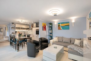 En moderne stue med sofa, sofabord og stole, et spiseområde med bord og stole, samt et køkken i baggrunden.