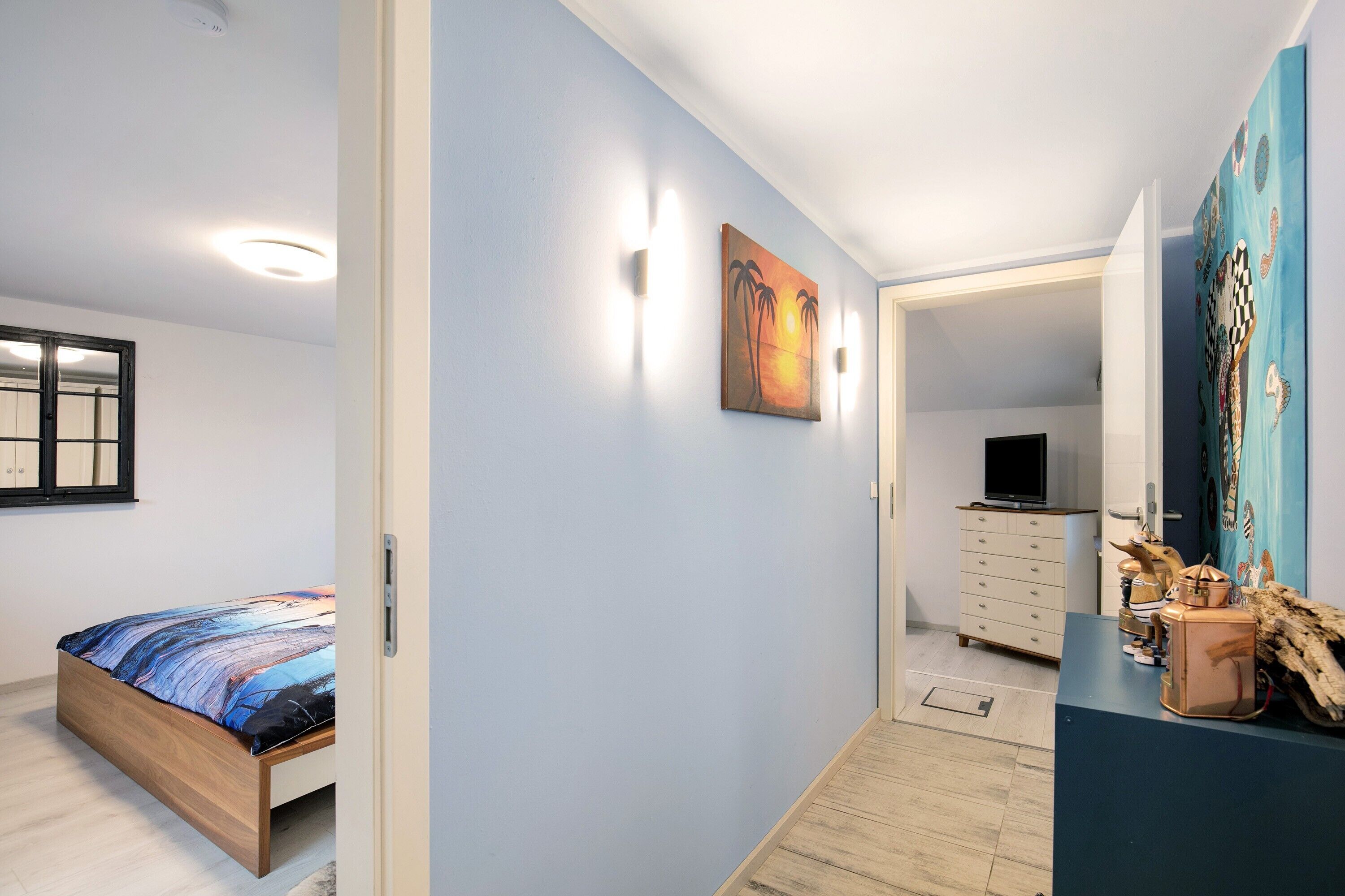 A bedroom with a bed, a painting, a dresser, and a television in the adjacent room.