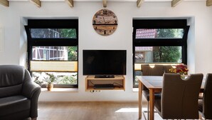 Una sala de estar con televisión, vigas de madera, ventanas grandes, un sofá y una mesa de comedor.