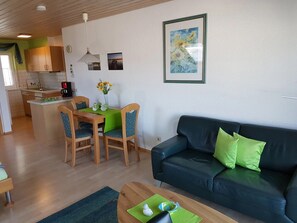 A living room with a sofa and chairs, a dining table with chairs, a kitchen area, and a framed picture on the wall.