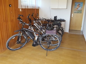 Una stanza con pavimento in legno, una presa elettrica a parete, una porta con un poster e un porta biciclette con diverse biciclette.