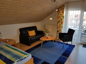 A room with a black leather sofa, a wooden coffee table, a black chair, a bed with a striped blanket, and a window with sheer curtains.