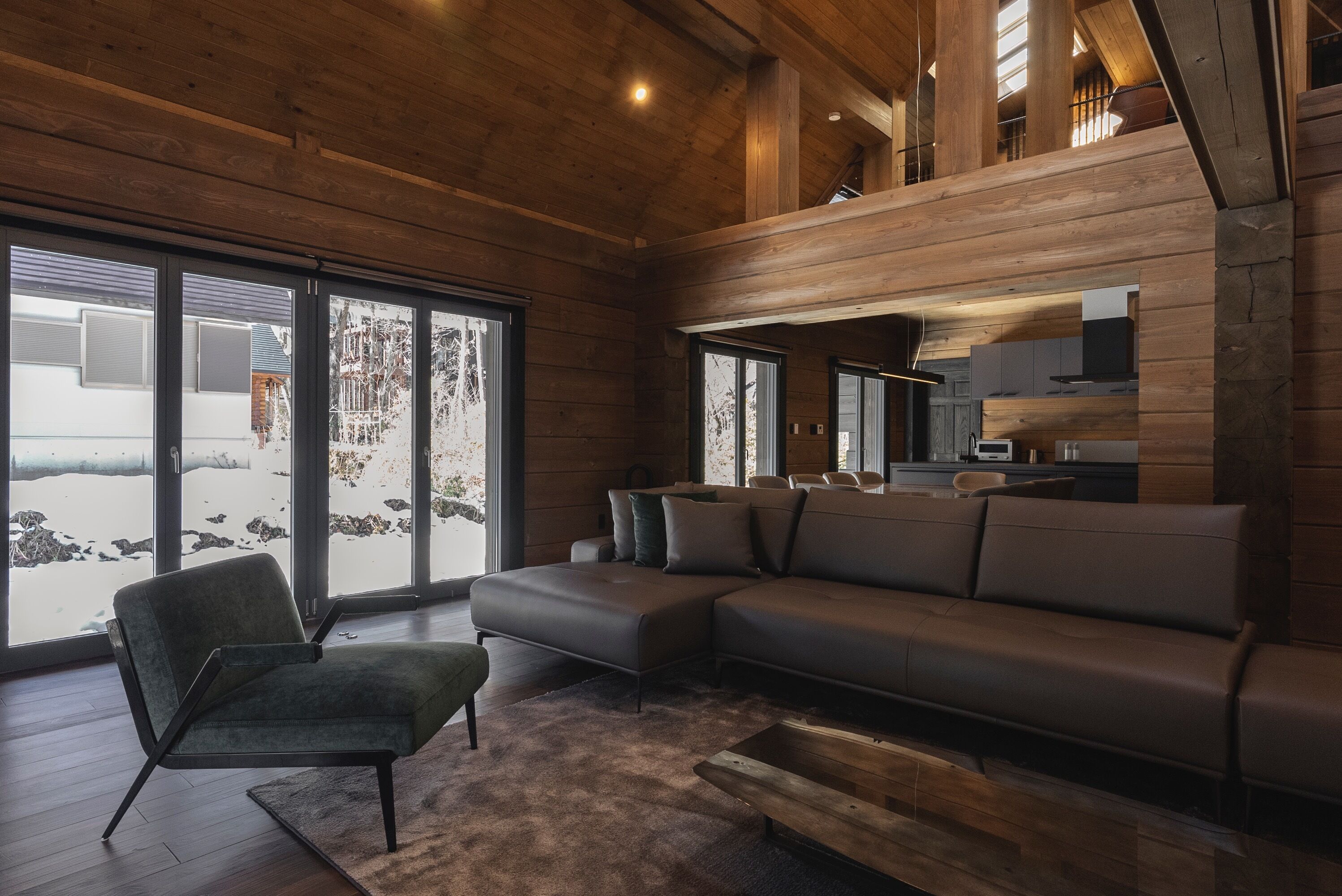 Ein modernes Wohnzimmer mit einer Eckcouch, einem grünen Sessel und einem Couchtisch aus Holz. Große Fenster bieten Blick auf eine verschneite Landschaft.