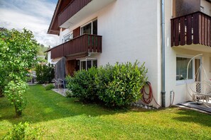 A two-story building with a balcony, a garden with a lawn and bushes, and a hose on the ground.