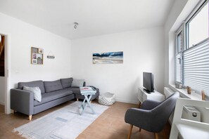 Un salon moderne avec un canapé gris, une petite table, un téléviseur et une fenêtre équipée de stores.