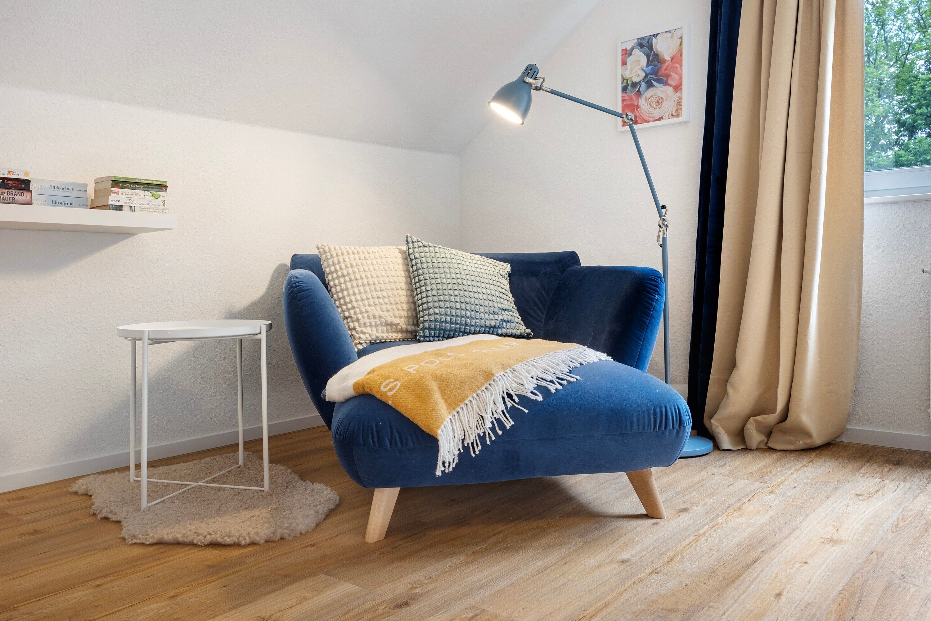 Un sillón azul con una manta, una mesita auxiliar blanca, una lámpara de pie, una estantería y una ventana con cortinas.