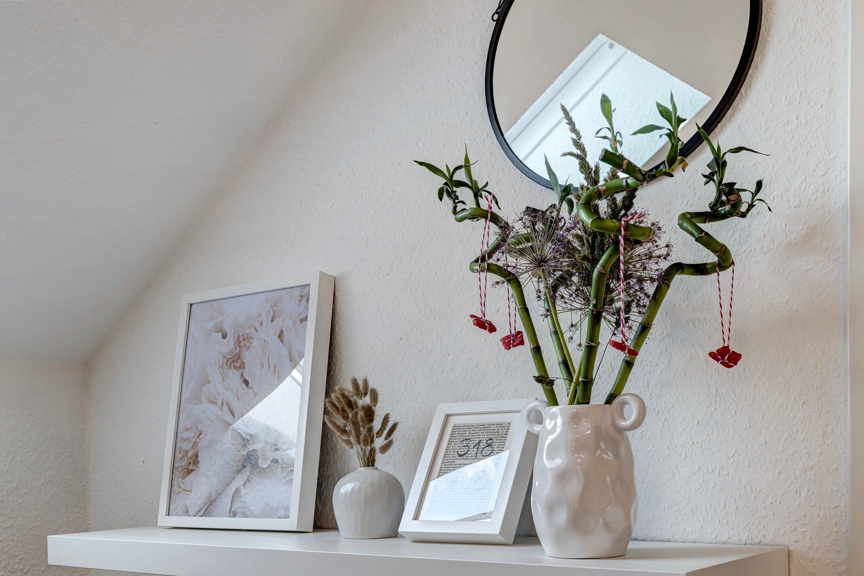 Una estantería blanca con una obra de arte enmarcada, un pequeño jarrón con plantas secas y una jarra de cerámica blanca con un arreglo floral.