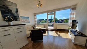 A modern living room with a sofa, a black chair, a dining area with a TV, and a large window with a view of houses. - Apartment 'Penthouse Laboe' with Sea View, Private Terrace and Wi-Fi (Laboe)