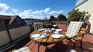 A rooftop terrace with a round wooden table set for four, surrounded by checkered chairs, and a view of buildings in the background. - Apartment 'Penthouse Laboe' with Sea View, Private Terrace and Wi-Fi (Laboe)