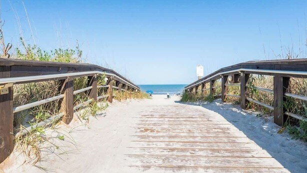 Un paseo marítimo de madera que conduce a la playa, bordeado por barandas de madera y vegetación.