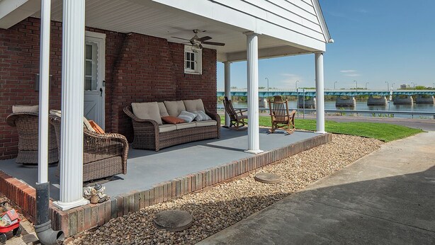A covered porch with a brick wall, a wicker sofa, and a view of a bridge over water. - River House - Harrisburg City Skyline Views with Game Room and Covered Porch (Wormleysburg)