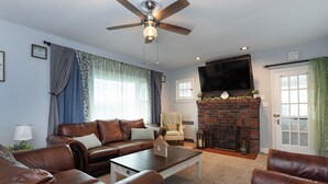 A living room with a brown leather sofa, a coffee table, a fireplace, and a ceiling fan. - River House - Harrisburg City Skyline Views with Game Room and Covered Porch (Wormleysburg)