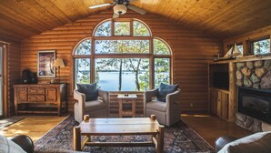 A cozy cabin interior with a large arched window, a fireplace, a wooden dining table, and comfortable seating. - Lakefront Cabin on LCO - The Sunset Escape (Hayward)