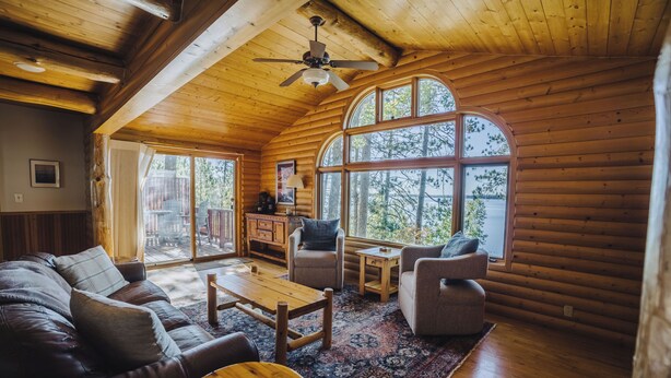 A cozy log cabin interior with a wooden ceiling, a ceiling fan, a large arched window, a leather sofa, a coffee table, and a small side table. - Lakefront Cabin on LCO - The Sunset Escape (Hayward)