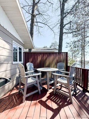 A covered deck with a table and four Adirondack chairs, overlooking a body of water. - Lakefront Cabin on LCO - The Sunset Escape (Hayward)