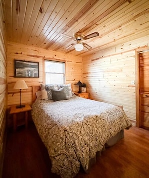 A cozy bedroom with a wooden ceiling, a bed with patterned bedding, a nightstand with a lamp, and a window with blinds. - Lakefront Cabin on LCO - The Sunset Escape (Hayward)