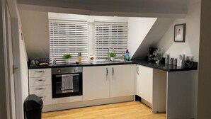 Una cocina moderna con armarios blancos, encimera negra y horno empotrado. Hay dos ventanas con persianas, un lavabo y varios utensilios de cocina sobre la encimera.