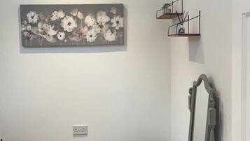 Une pièce comprenant un pouf capitonné gris, une commode en bois avec un miroir, un tableau floral gris accroché au mur et une petite étagère avec des plantes.