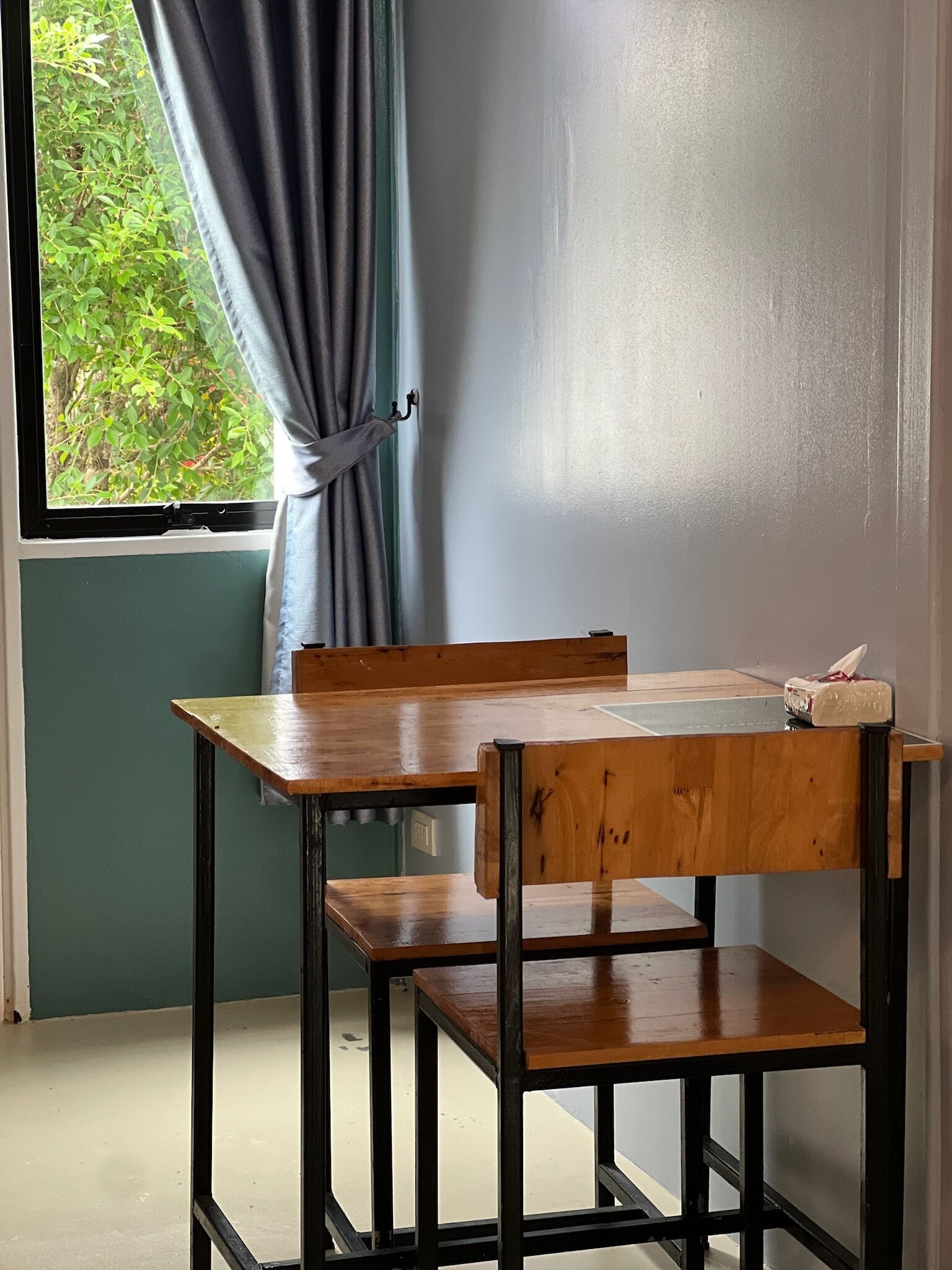 Une petite pièce simple, avec une table et une chaise en bois, une fenêtre avec un rideau et une vue sur la verdure à l’extérieur.
