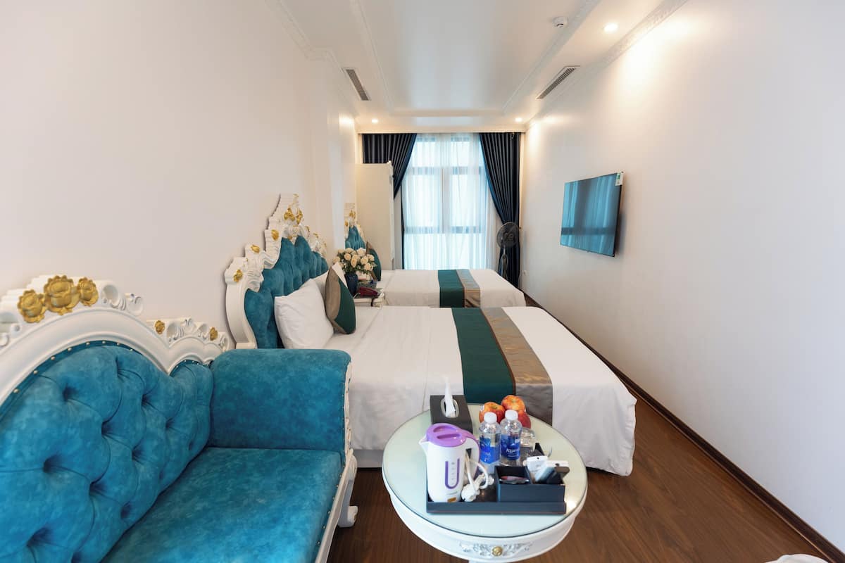 A hotel room with a blue sofa, two beds, a round coffee table with amenities, and a large window with curtains.