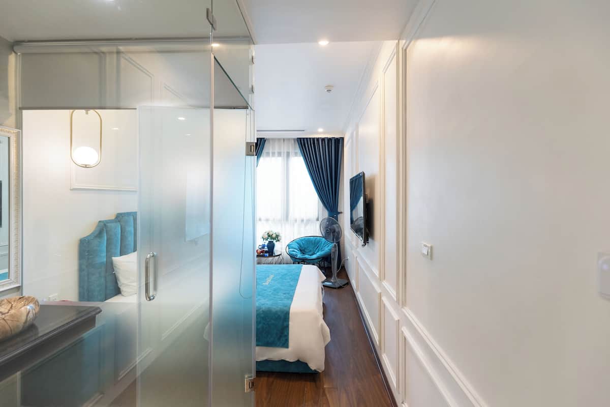 A modern bedroom with a glass door leading to a bathroom, a bed with a blue and white bedspread, and a flat-screen TV mounted on the wall.