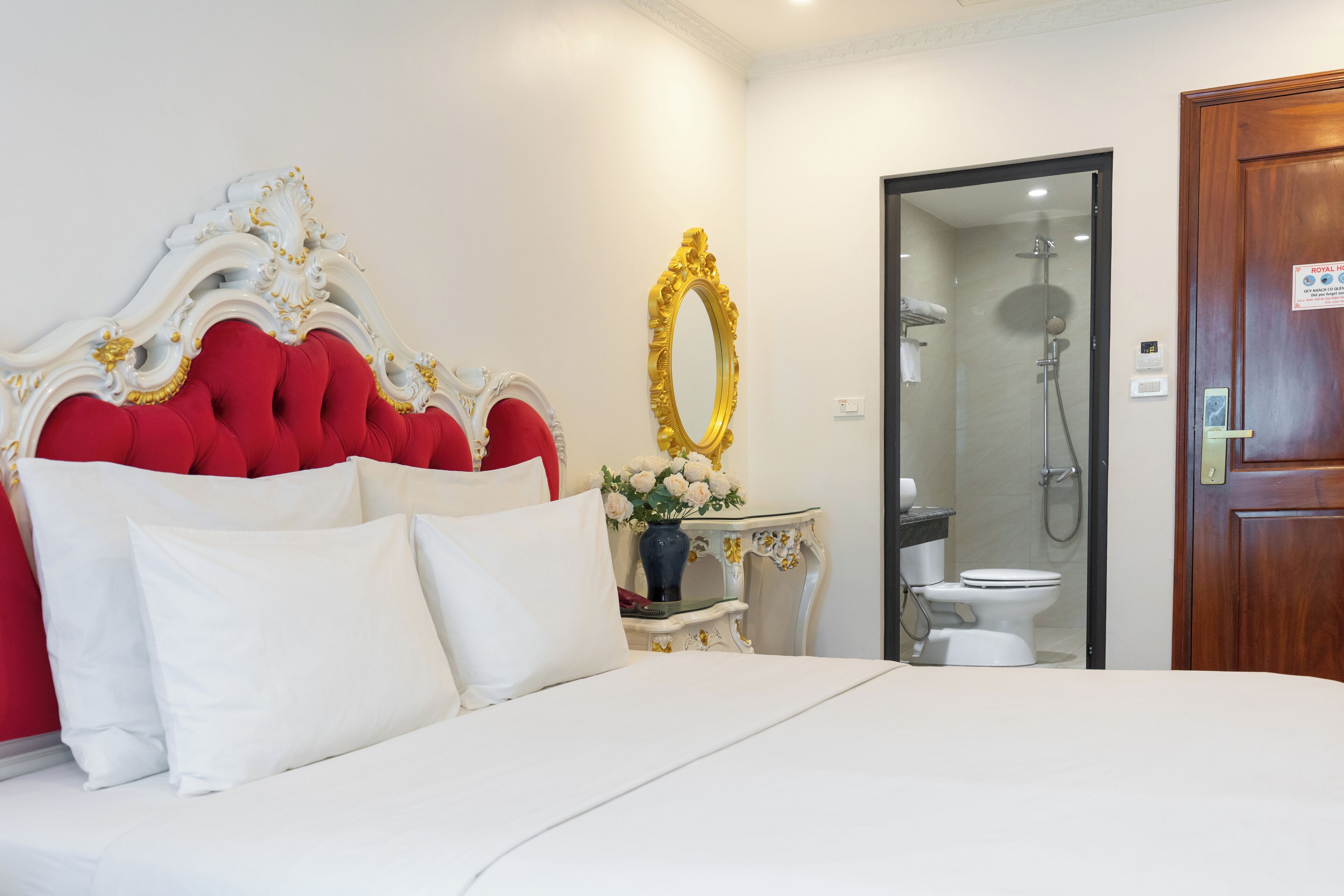 A hotel room with a large bed, a red upholstered headboard, a white bedside table with a vase of flowers, a gold-framed mirror, and a wooden door.