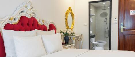 A hotel room with a large bed, a red upholstered headboard, a white bedside table with a vase of flowers, a gold-framed mirror, and a wooden door.