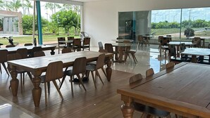 Un vaste espace de restauration intérieur, doté de grandes tables et de chaises en bois, de grandes fenêtres offrant une vue sur la verdure et un bâtiment situé à l’extérieur.