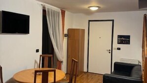 Une pièce comprenant une table de salle à manger ronde en bois, quatre chaises en bois, un canapé noir, un téléviseur fixé au mur et une porte ornée d’une affiche.