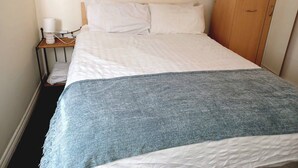 A single bed with a white and blue blanket, a bedside table with a lamp, and a wooden wardrobe.