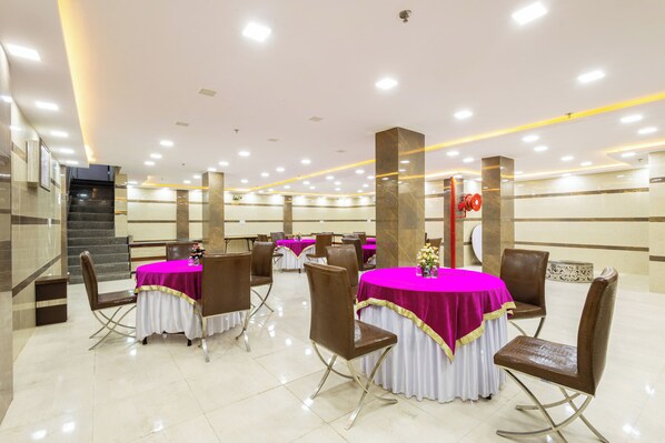 A spacious banquet hall with multiple round tables covered with white and pink tablecloths, brown chairs, and a central staircase.