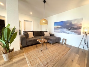 Ein Wohnzimmer mit grauem Sofa, einem runden Couchtisch, einem großen Webteppich, einer hängenden Leuchte und einem Wandbild mit Strandmotiv.