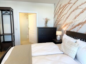 Un dormitorio con cama, cómoda, mesita de noche, lámpara y una puerta que da a otra habitación.
