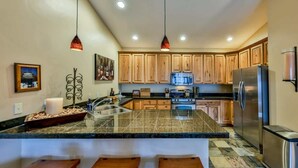 A modern kitchen with wooden cabinets, granite countertops, a stainless steel refrigerator, and pendant lighting.