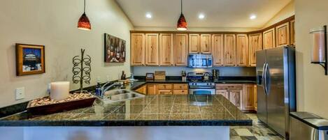 A modern kitchen with wooden cabinets, granite countertops, a stainless steel refrigerator, and pendant lighting.