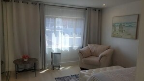 Habitación de hotel con un ventanal grande, un radiador, un sillón color beige, una mesita con un jarrón y un cuadro en la pared.