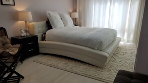 Un dormitorio con una cama curva, dos lámparas de noche, una mesita de noche, una alfombra mullida y una silla.