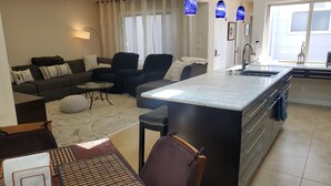 Un salón moderno con un sofá, sillas, una mesa de centro y un comedor con mesa y sillas. Hay una cocina con un fregadero y armarios.